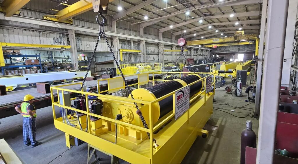 Worker guiding overhead crane lifting 45-ton hoist drum assembly in manufacturing facility.