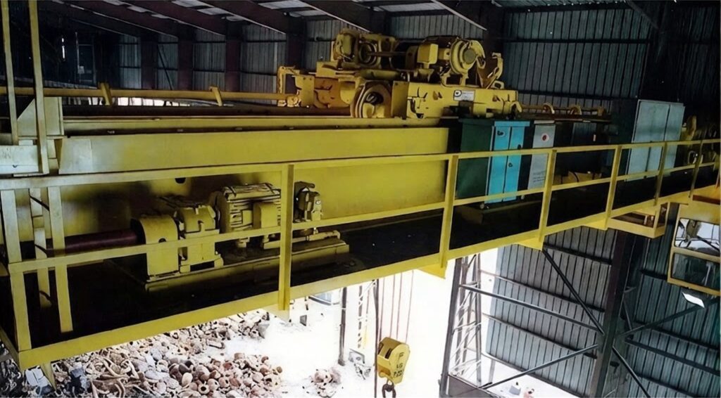 Yellow double girder overhead crane with walkway above scrap metal pile.